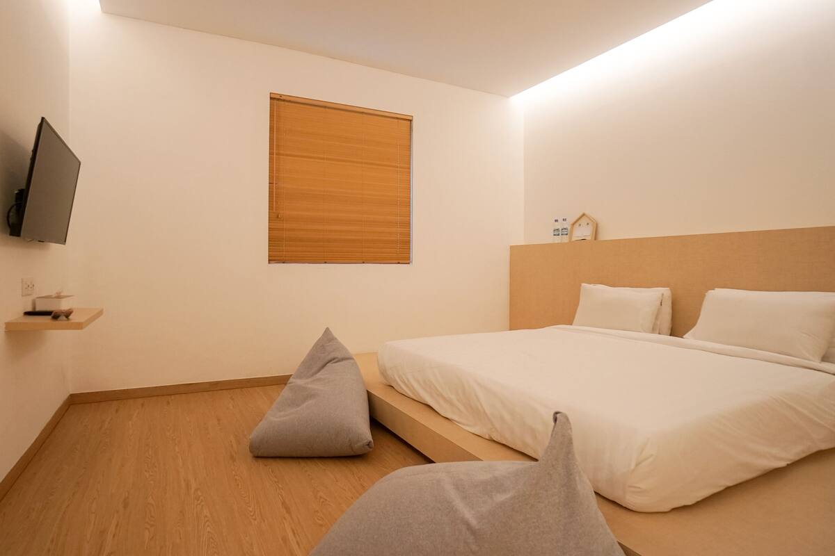 A bedroom with a bed, a television mounted on the wall, a window with blinds, and a wooden floor.