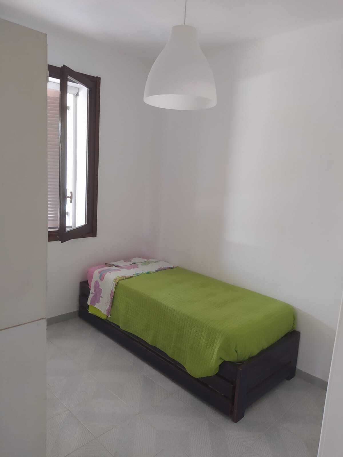 A small, single-bed room with a window, a hanging light fixture, and a door.