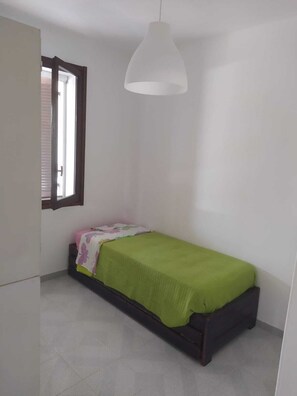 Quarto pequeno com cama de solteiro, janela, luminária pendente e porta.