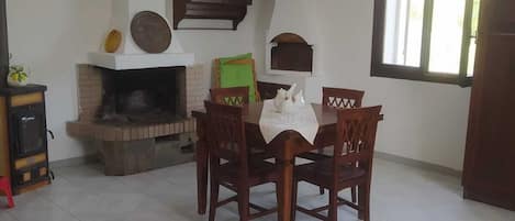 A dining area with a brick fireplace, a wooden table with chairs, a hanging light fixture, and a window with blinds.