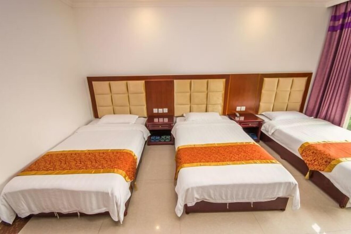 A hotel room with four single beds, each with a white bedspread and an orange decorative border. The room has a wooden headboard with a tufted design and a neutral-colored wall.