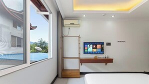 A hotel room with a bed, a TV mounted on the wall, a small wooden shelf, and a window with a view of a building and greenery. - Mingxiuya Leisure Villa (Emeishan City)