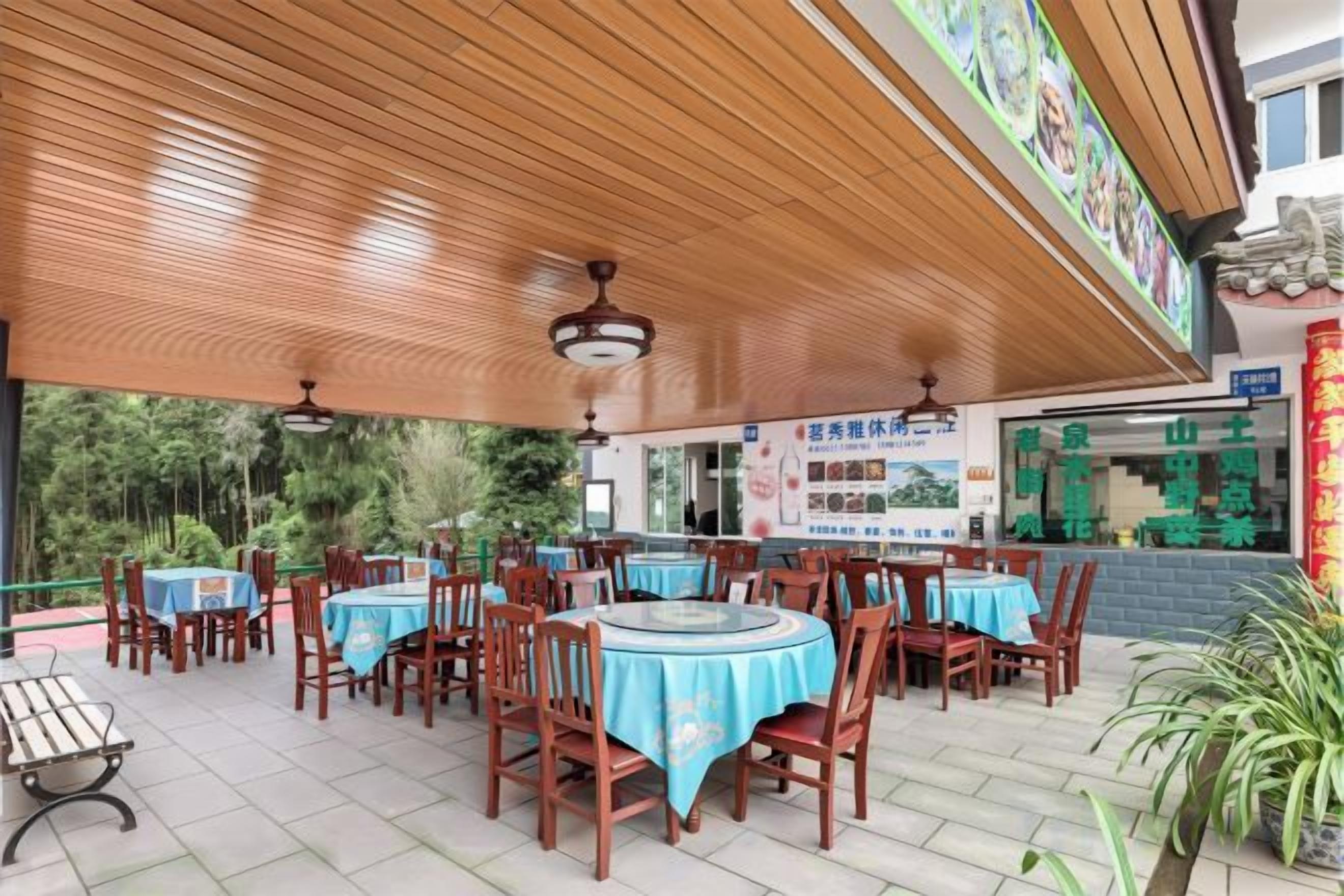 An outdoor dining area with round tables and chairs, covered by a wooden roof, and a signboard in the background.