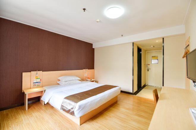 A hotel room with a wooden floor, a bed with white bedding, a nightstand, a desk, and a television.