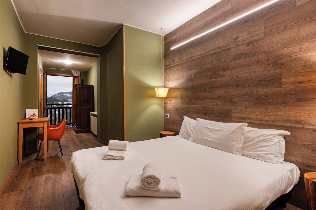 Une chambre d’hôtel avec un mur d’accent en bois, un lit avec du linge de maison blanc, un bureau avec une chaise et une vue sur les montagnes par une porte ouverte.