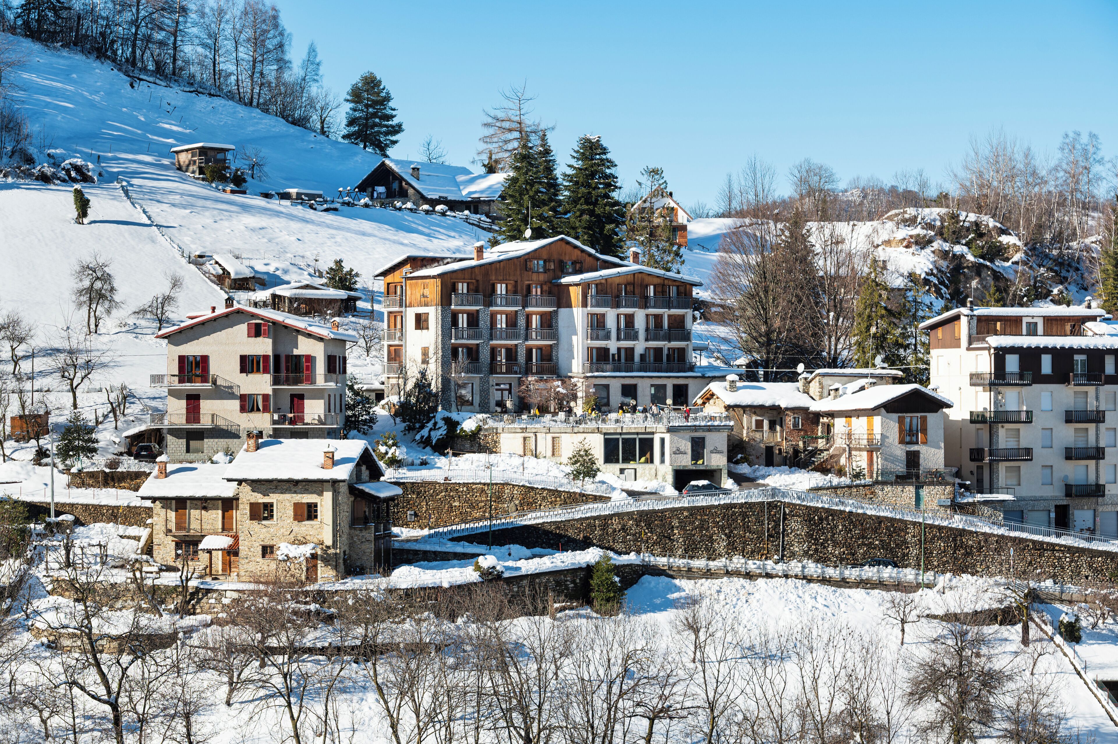 Ein verschneites Bergdorf mit mehrstöckigen Gebäuden, darunter ein Hotel mit Balkonen und ein Steinhaus mit rotem Dach.
