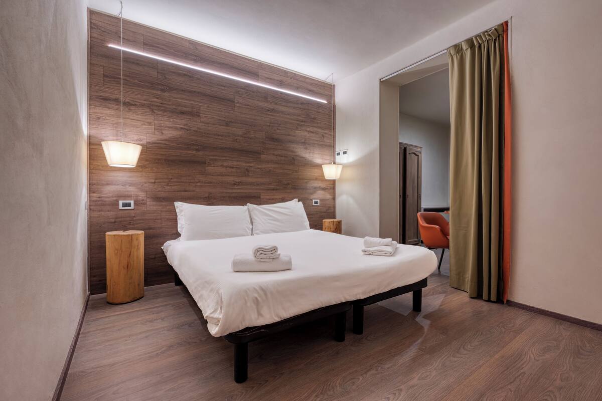 Une chambre d’hôtel moderne avec une tête de lit en bois, un grand lit avec du linge de lit blanc, deux lampes de chevet et une table de chevet en bois.