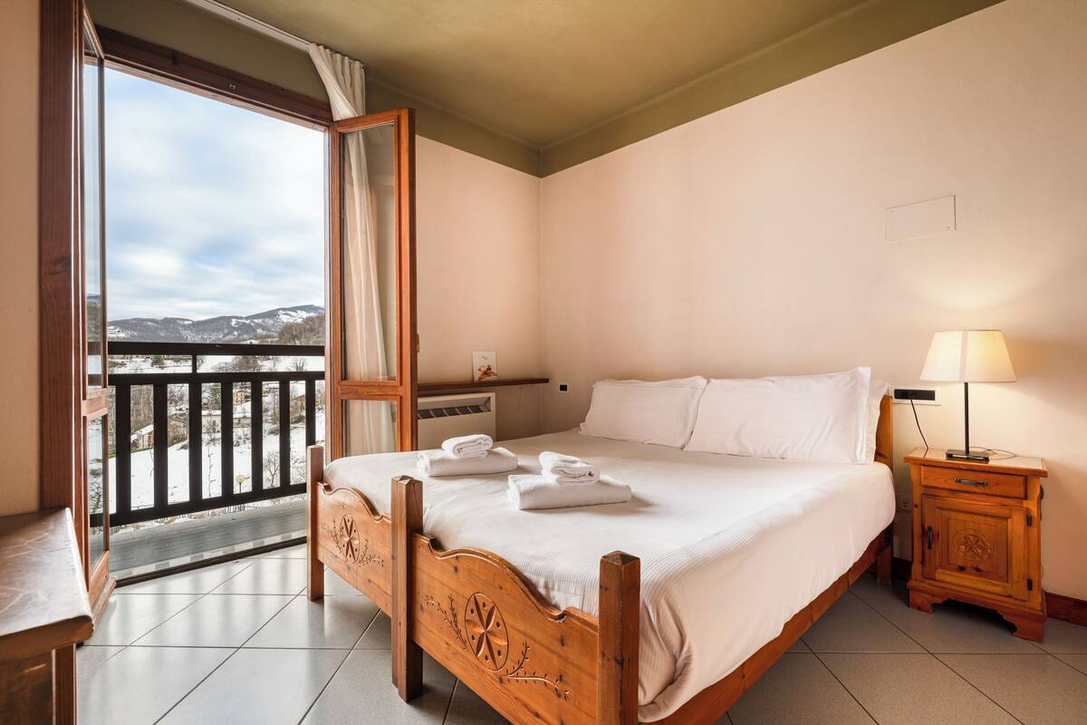 Une chambre avec un lit en bois, une table de chevet avec une lampe, et une vue sur des montagnes enneigées par la porte d’un balcon.
