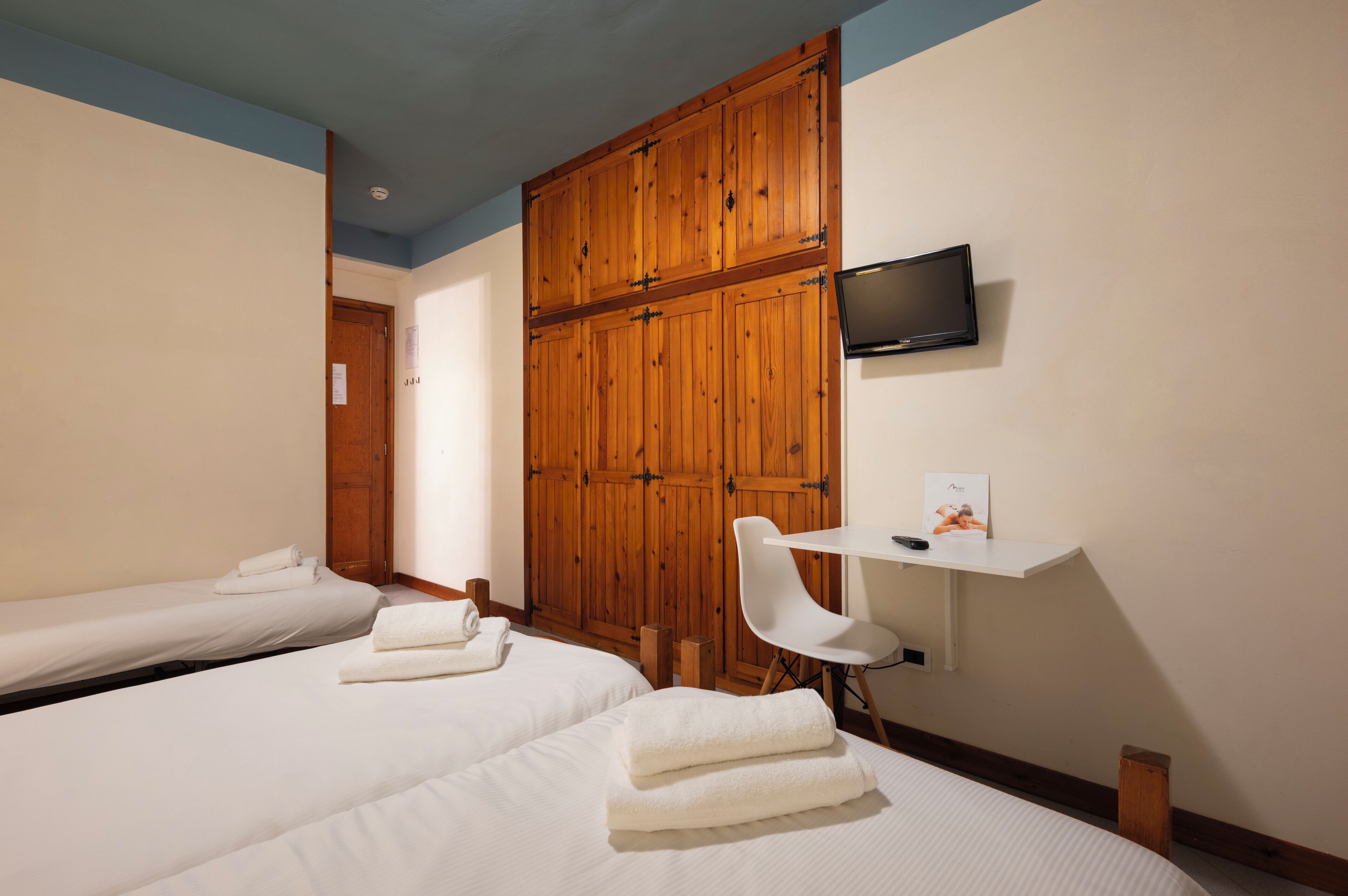 A hotel room with two beds, a wooden wardrobe, a TV mounted on the wall, and a small desk with a chair.