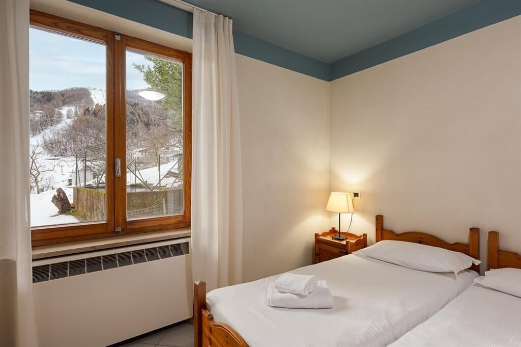 Habitación de hotel con una ventana grande, una cama, una mesita de noche con lámpara y vista a un paisaje nevado.