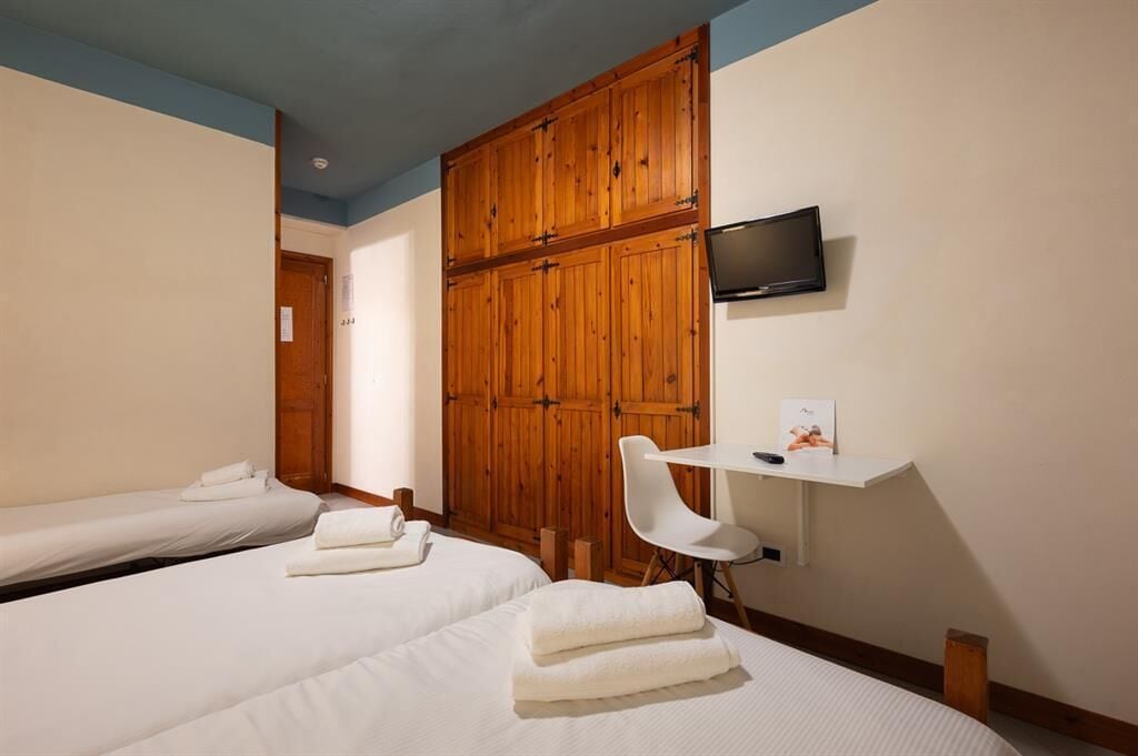 Quarto de hotel com duas camas, um guarda-roupa de madeira, uma televisão e uma mesa de trabalho com cadeira.