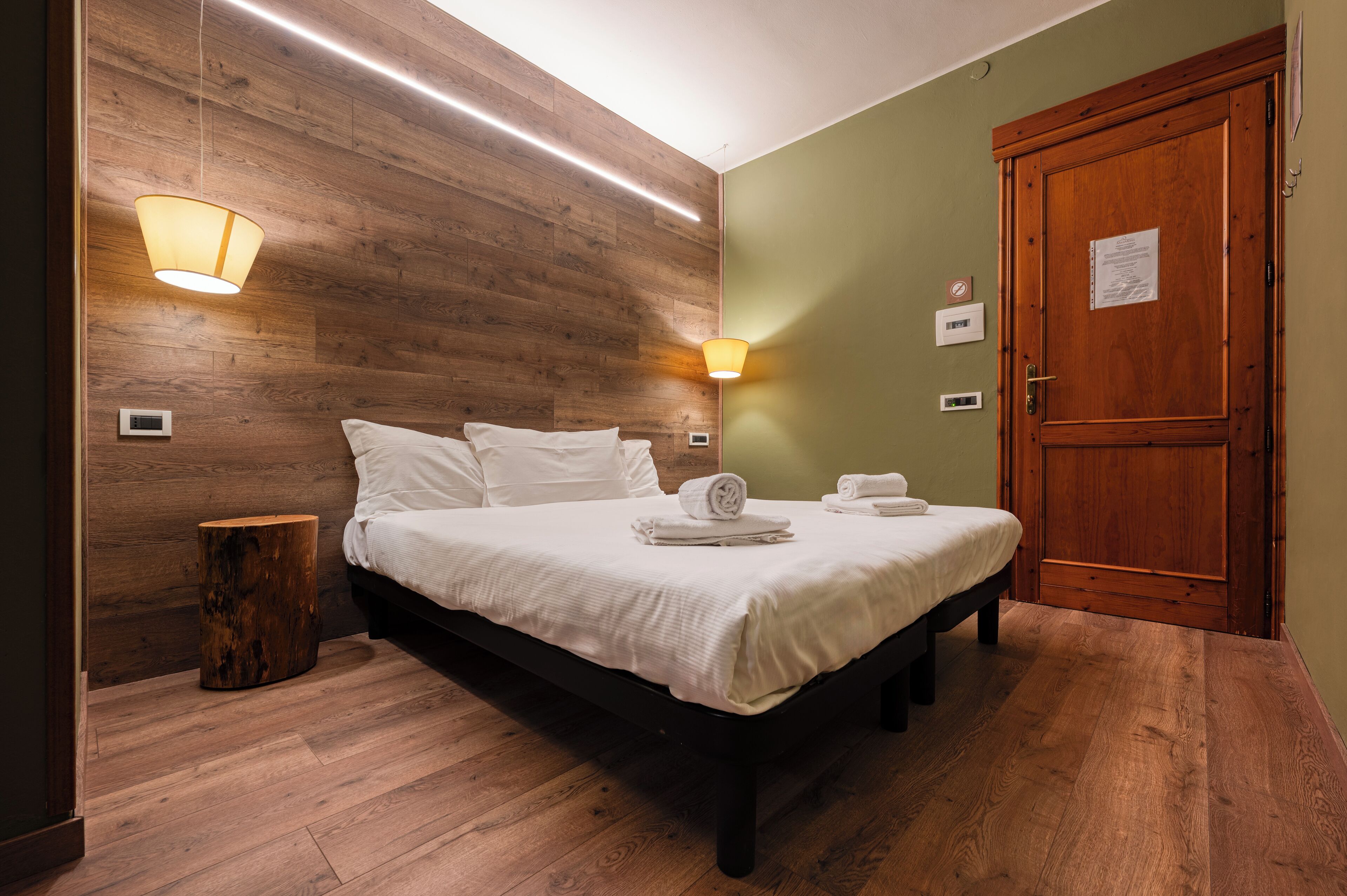 A neatly made bed with white linens, a wooden headboard, and a wooden floor. There are two wall-mounted light fixtures and a wooden door with a sign on it.