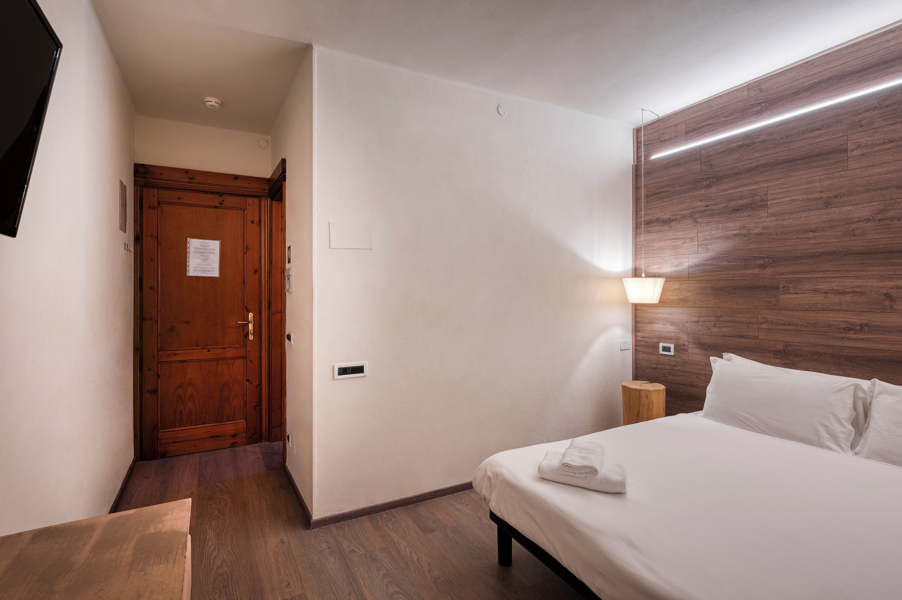 Une chambre d’hôtel avec une porte en bois, un lit avec du linge de maison blanc, une table de chevet et un luminaire fixé au mur.