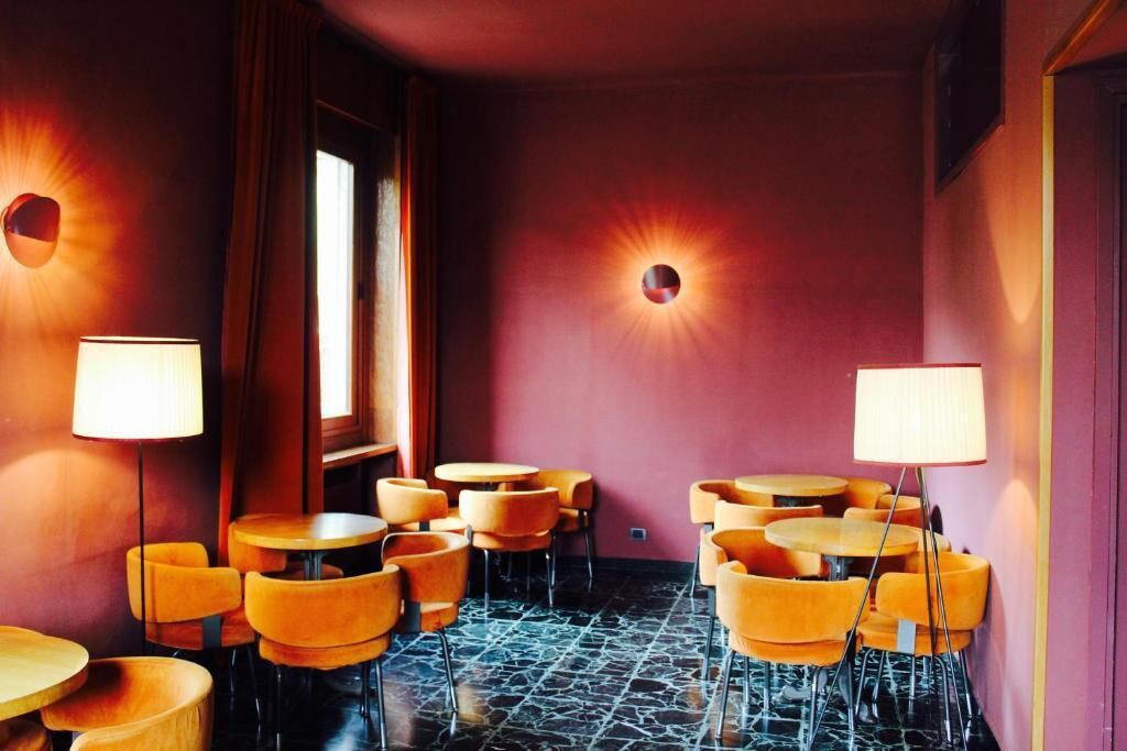A room with round tables and orange chairs, a marble floor, and a red wall with a circular light fixture.