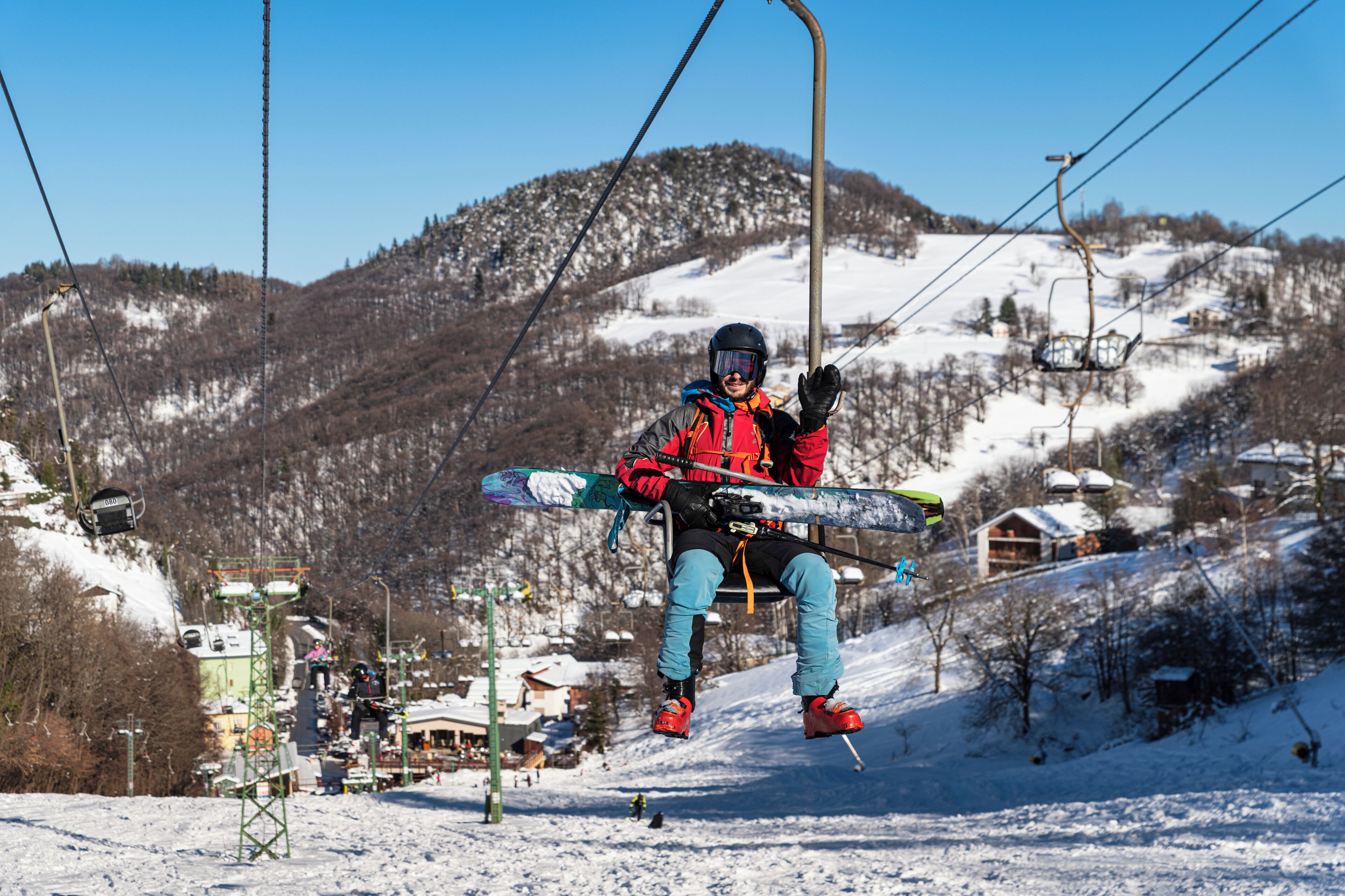 A person seated on a ski lift chair holding skis, with a snowy mountain landscape in the background.