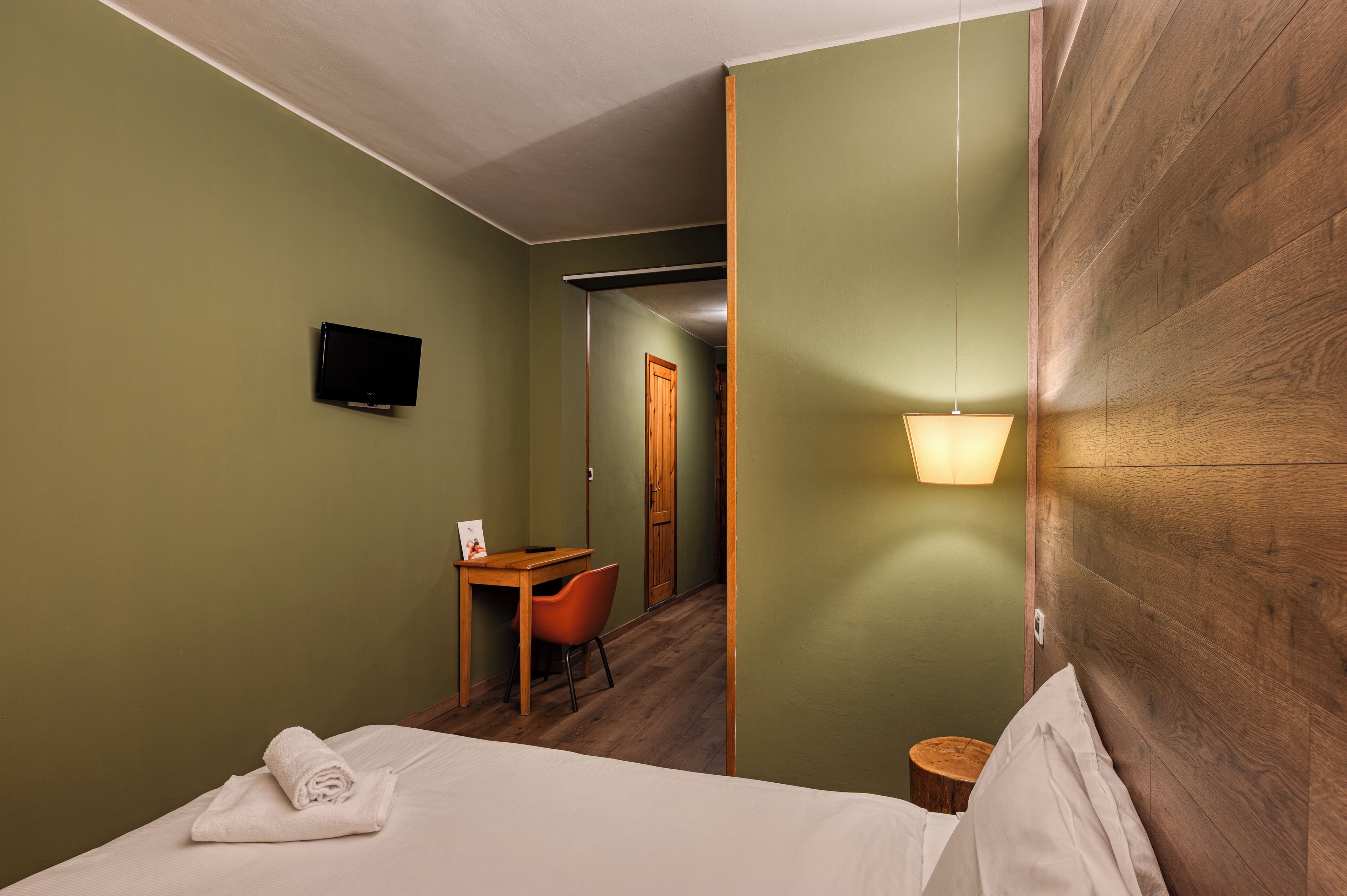 Quarto de hotel com cama, escrivaninha com cadeira, televisão e cabeceira de madeira.