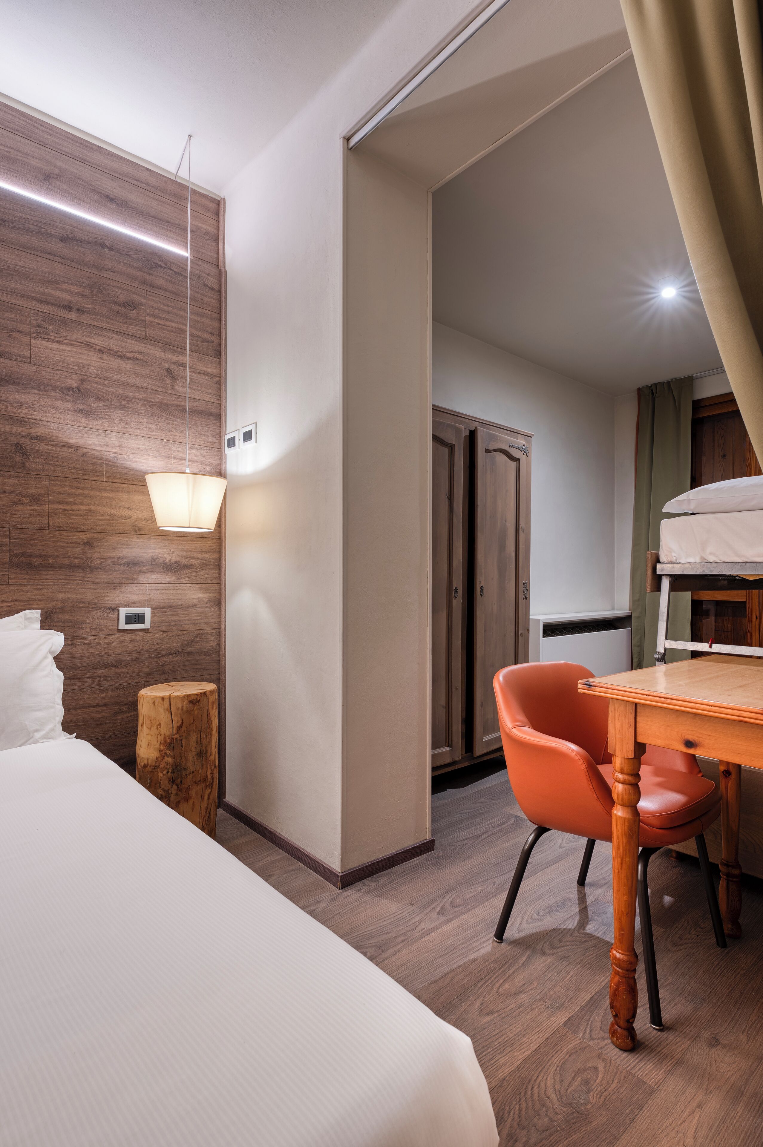 A hotel room with a bed, a wooden wall, a desk with a chair, a hanging light fixture, and a wardrobe.
