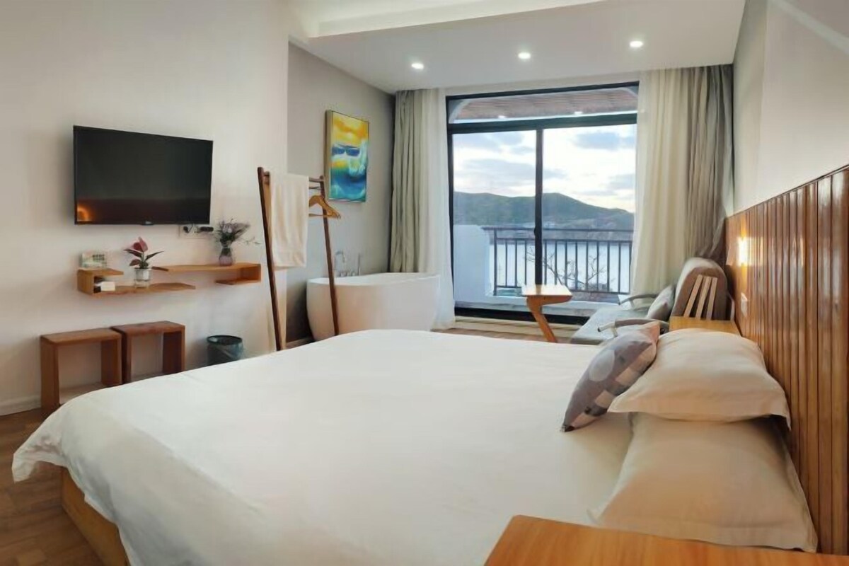 A hotel room with a large bed, a television, a window with a view of the sea, and a balcony.