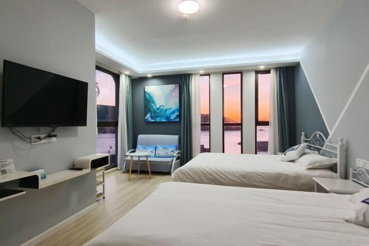 A hotel room with two beds, a TV, a desk, and a large window with a view of the sunset.