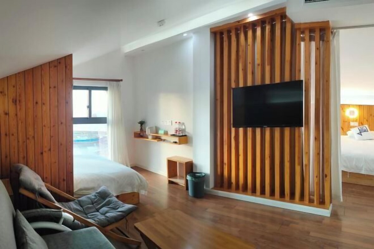 A modern room with a bed, a seating area with a chair and a small table, a television mounted on a wooden panel, and a window providing natural light.