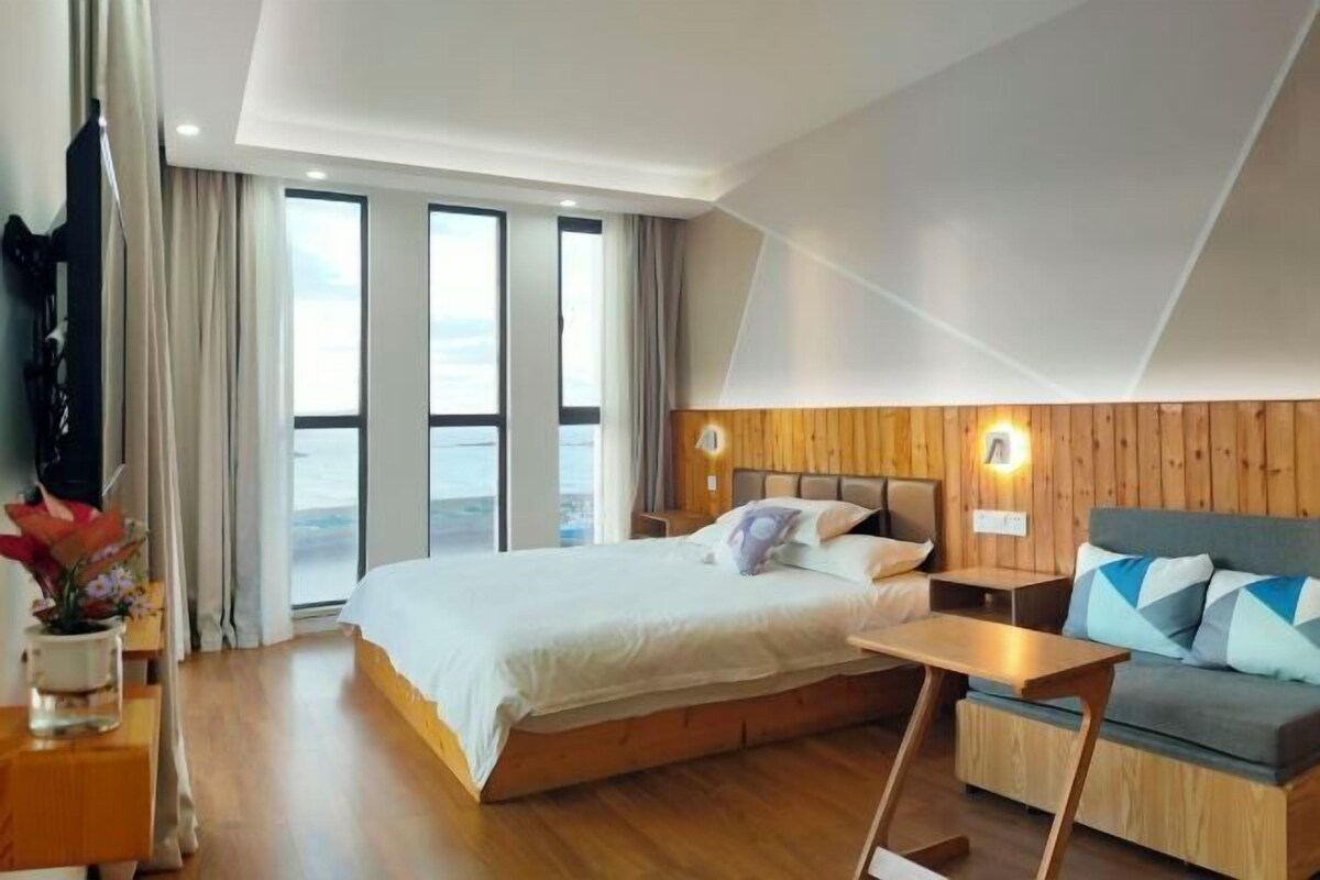 A modern hotel room with a large bed, a sofa, a small table, and a view of the outdoors through large windows.