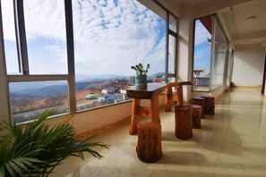 A spacious room with large windows offering a view of the landscape. There is a wooden table with stools and a potted plant. - Kunming Red Earth Summer Homestay (Kunming)
