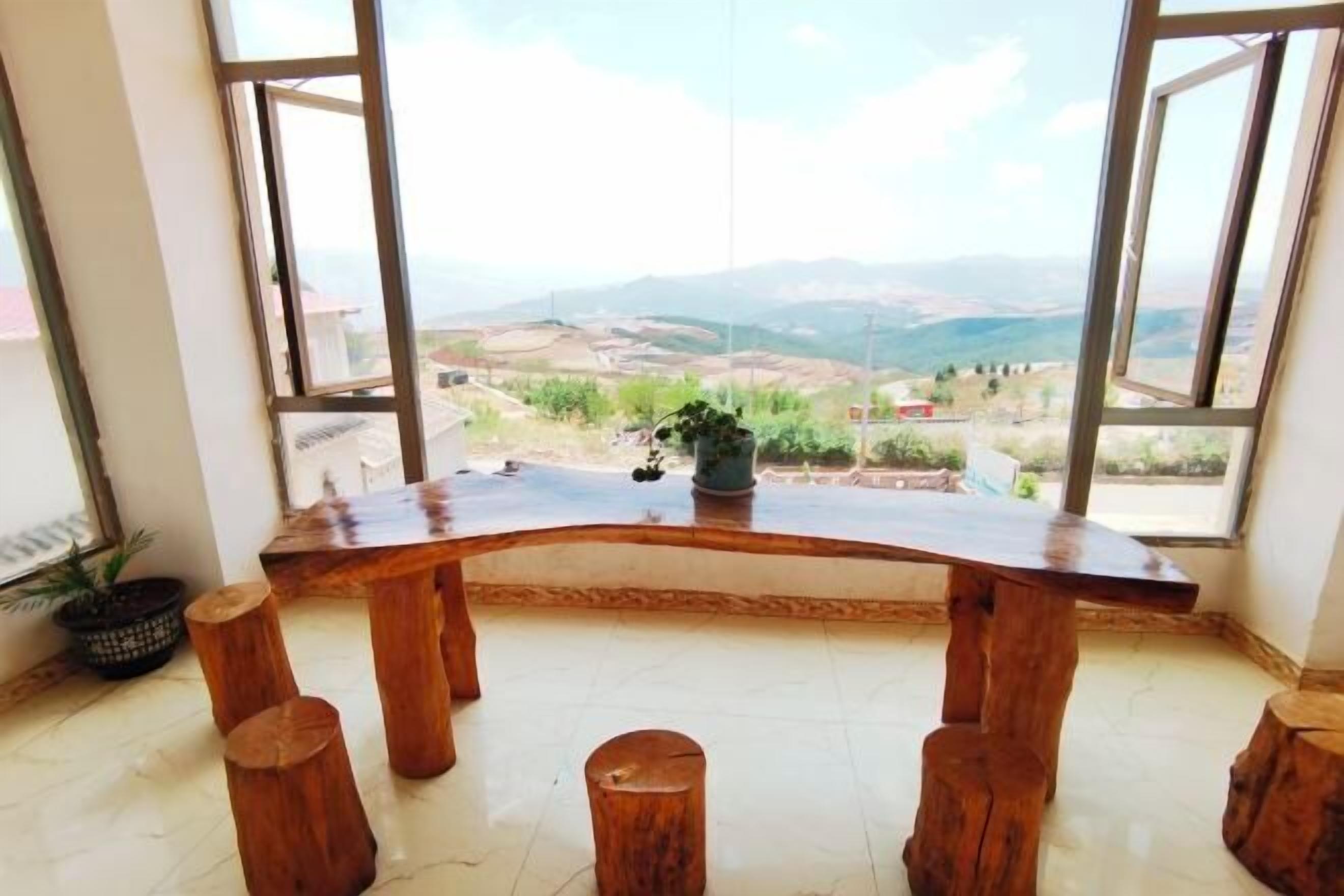 A wooden table with a curved top, surrounded by four wooden stools, set in a room with large windows offering a view of a landscape with hills and buildings.