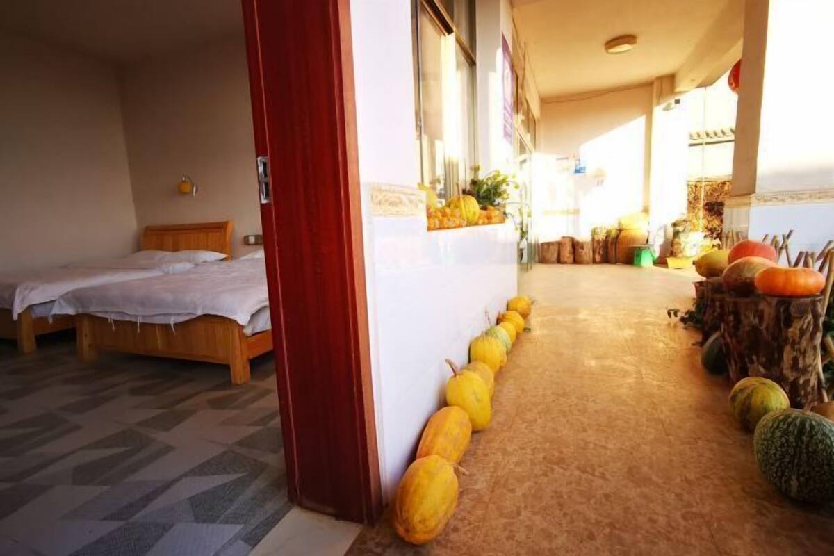 A hotel room with a bed, a wooden headboard, a white wall, and a hallway with pumpkins.