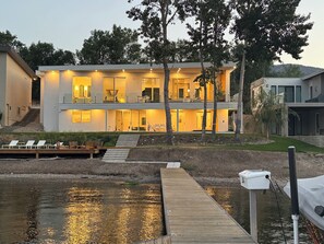 Une maison moderne à deux étages, dotée d’un quai s’avançant dans un plan d’eau, et entourée d’arbres et d’un paysage vallonné.