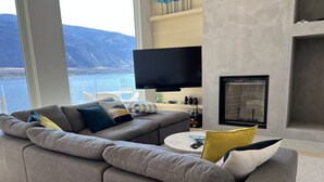 A modern living room with a grey sectional sofa, a flat-screen TV, a fireplace, and a view of a lake and mountains.