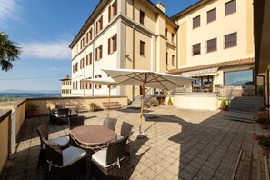 Eine Terrasse mit Korbmöbeln und einem großen Sonnenschirm, mit Blick auf ein Gebäude mit mehreren Fenstern und einer Treppe.