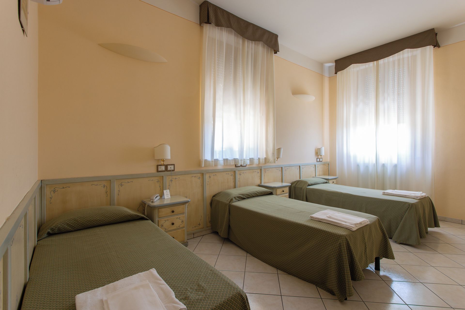 A hotel room with three single beds, a bedside table with a lamp, and a window with curtains.