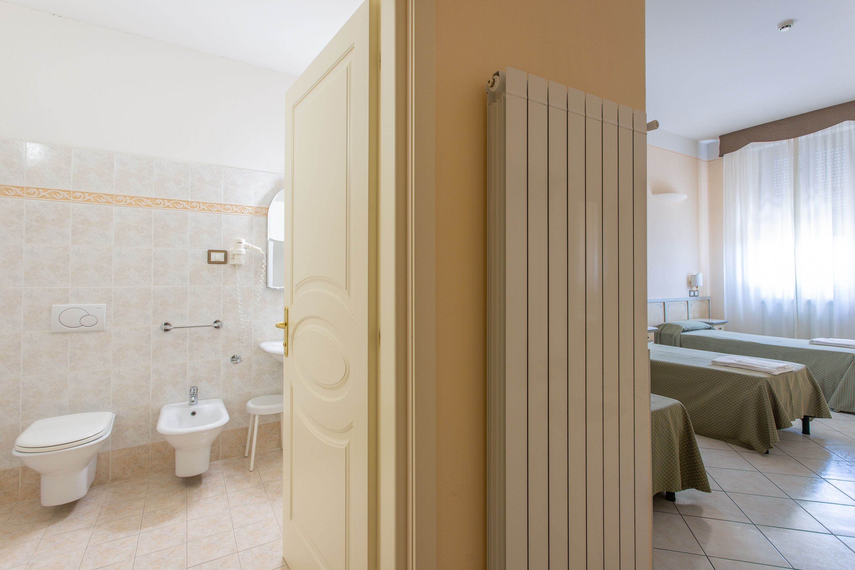 Ein Hotelbad mit Toilette, Bidet und Waschbecken. Im angrenzenden Zimmer befinden sich zwei Betten.