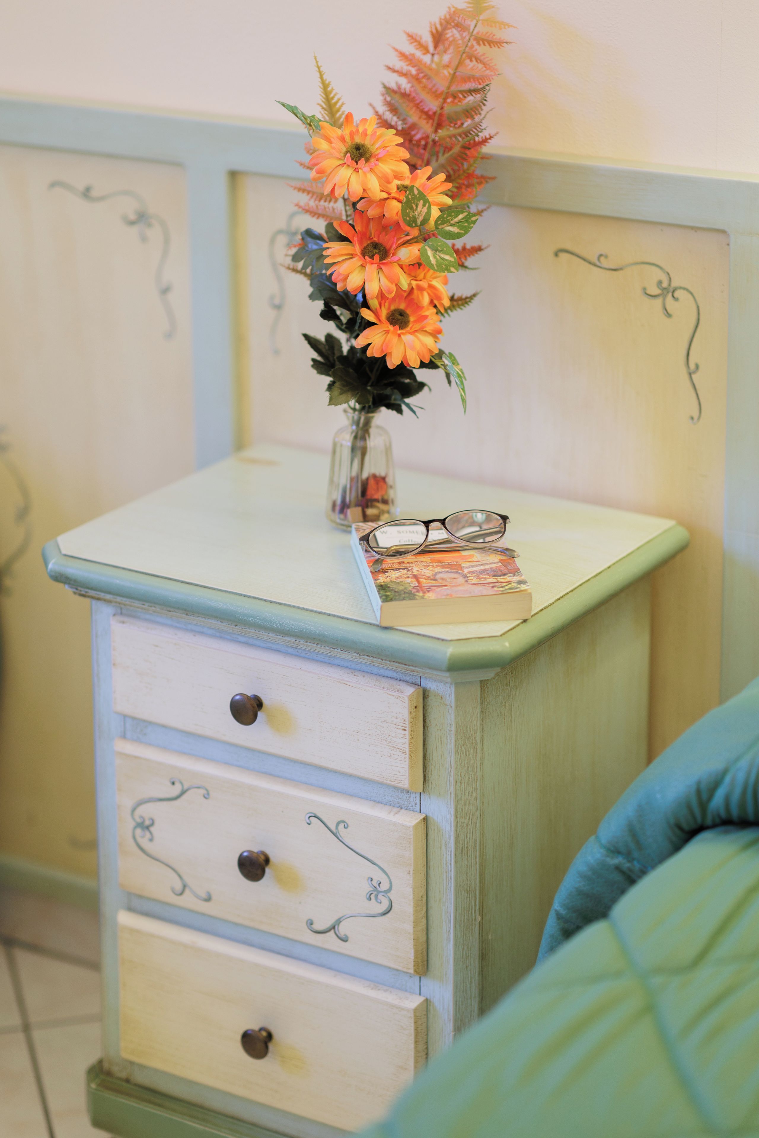 A vintage-style dresser with a vase of flowers on top, a pair of glasses, and a book.