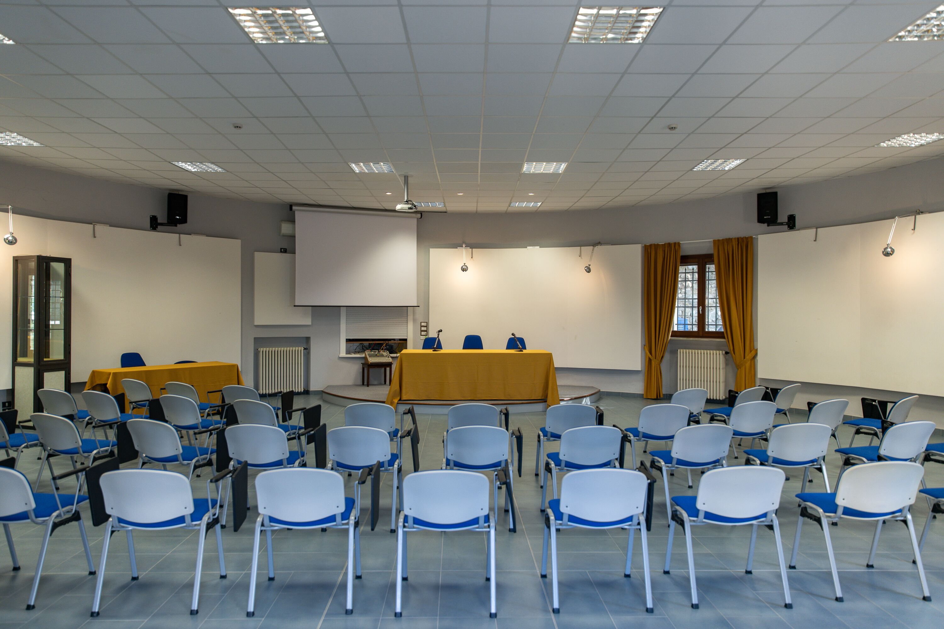 A conference room with rows of chairs, a table with a yellow tablecloth, a projector screen, and a speaker on the ceiling.