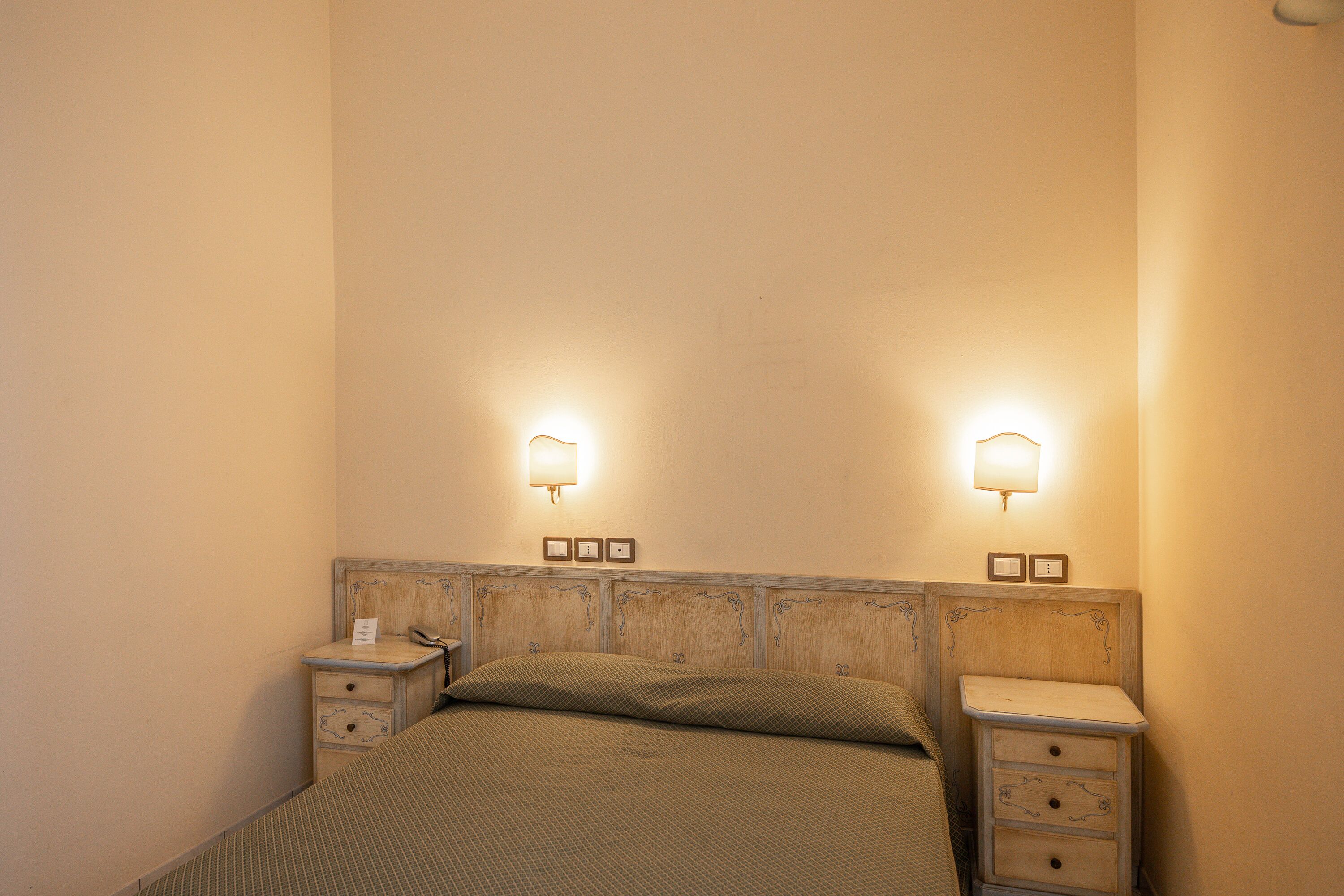 Een slaapkamer met een houten hoofdbord, twee nachtkastjes, een bed met een beddegoed met patroon, en wandlampen.