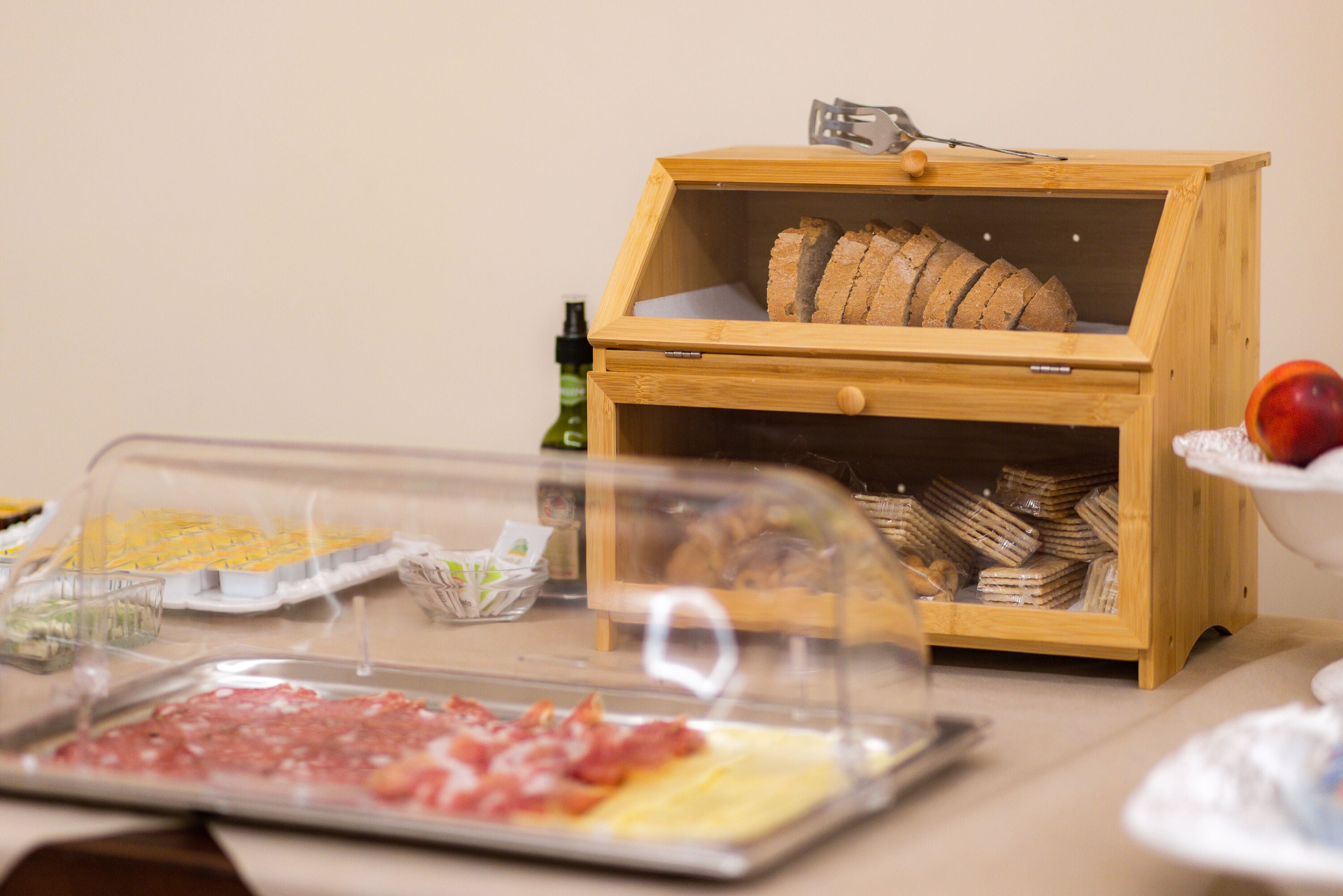 Um buffet de café da manhã com diversos alimentos, incluindo carnes, queijos e pães, expostos em um aparador de madeira e sob uma cobertura de vidro.