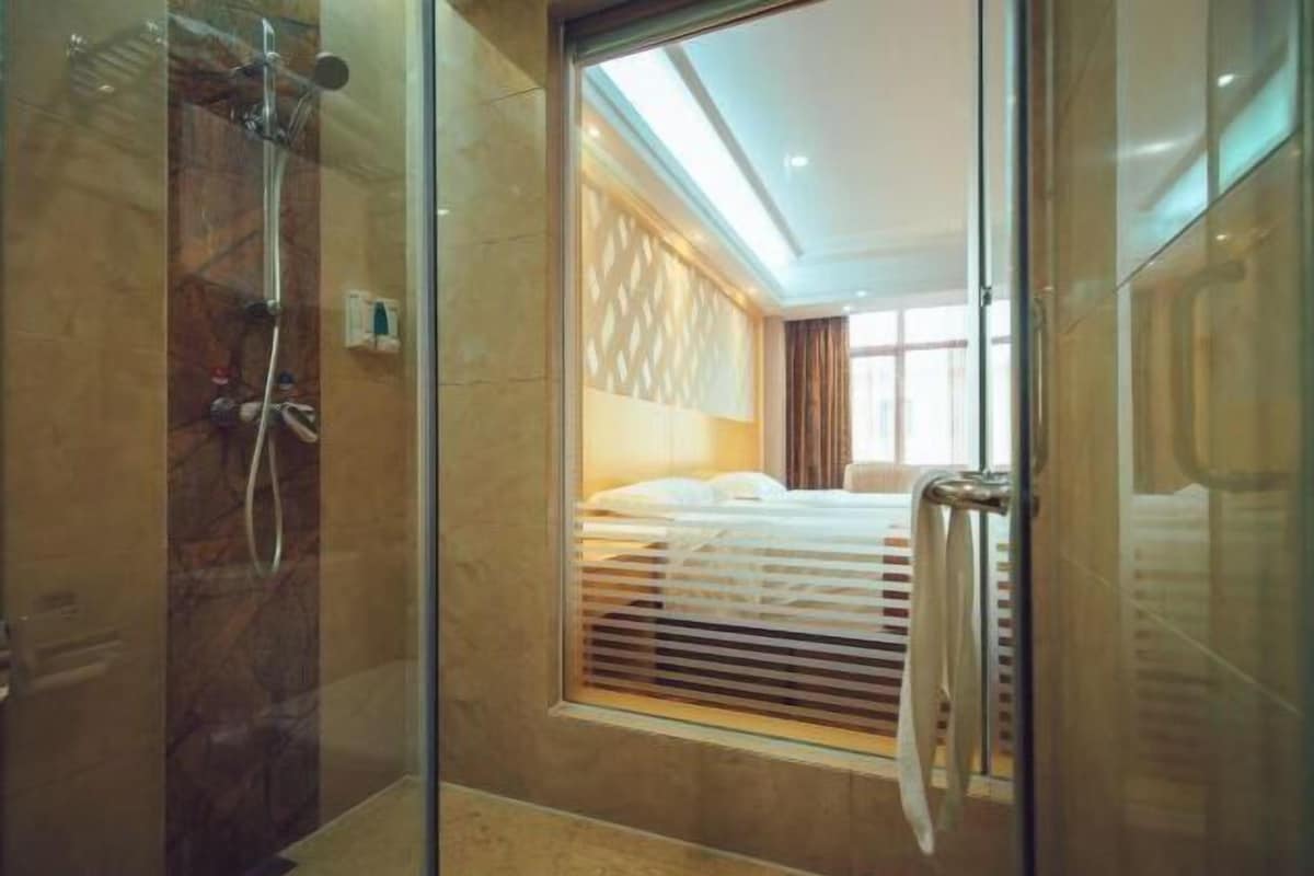 A modern hotel room with a glass shower enclosure, a bed with a headboard, and a window with blinds.