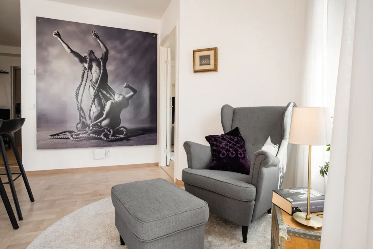 Un salon comprenant un grand tableau abstrait, un fauteuil gris agrémenté d’un coussin à motifs, un petit pouf et une table d’appoint avec une lampe et des livres.
