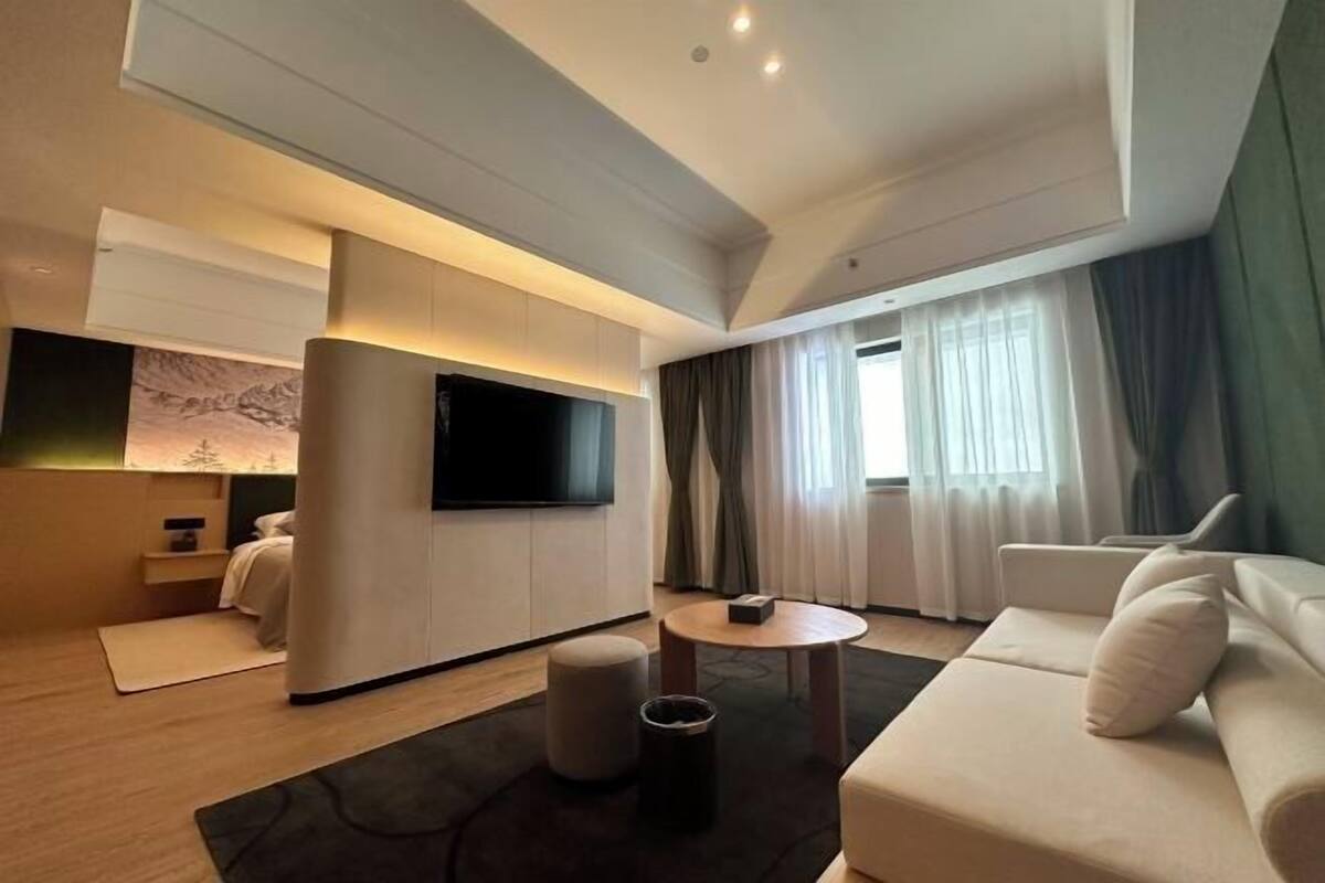 A modern hotel room with a sofa, a flat-screen TV, a round coffee table, and a large window with curtains.