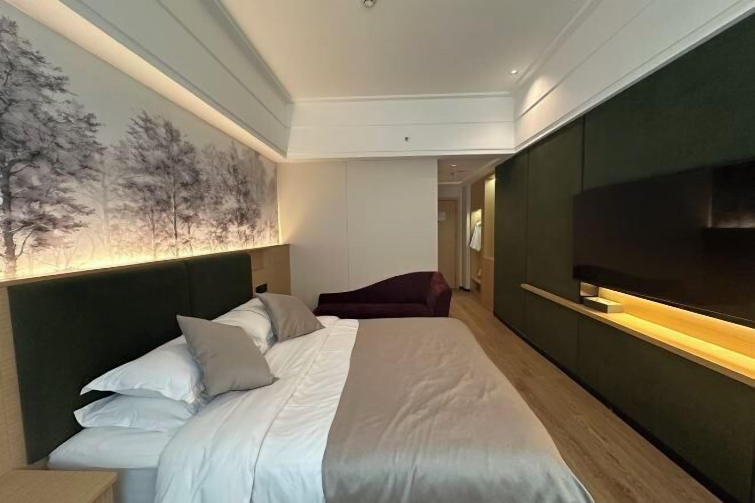 A modern hotel room with a large bed, a dark headboard, a wall-mounted TV, and a painting of trees.