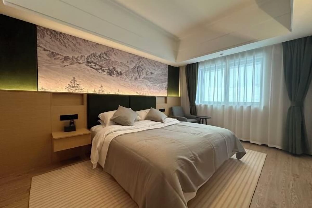 A modern hotel room with a large bed, a nightstand, a wall-mounted lamp, and a mountain landscape mural.