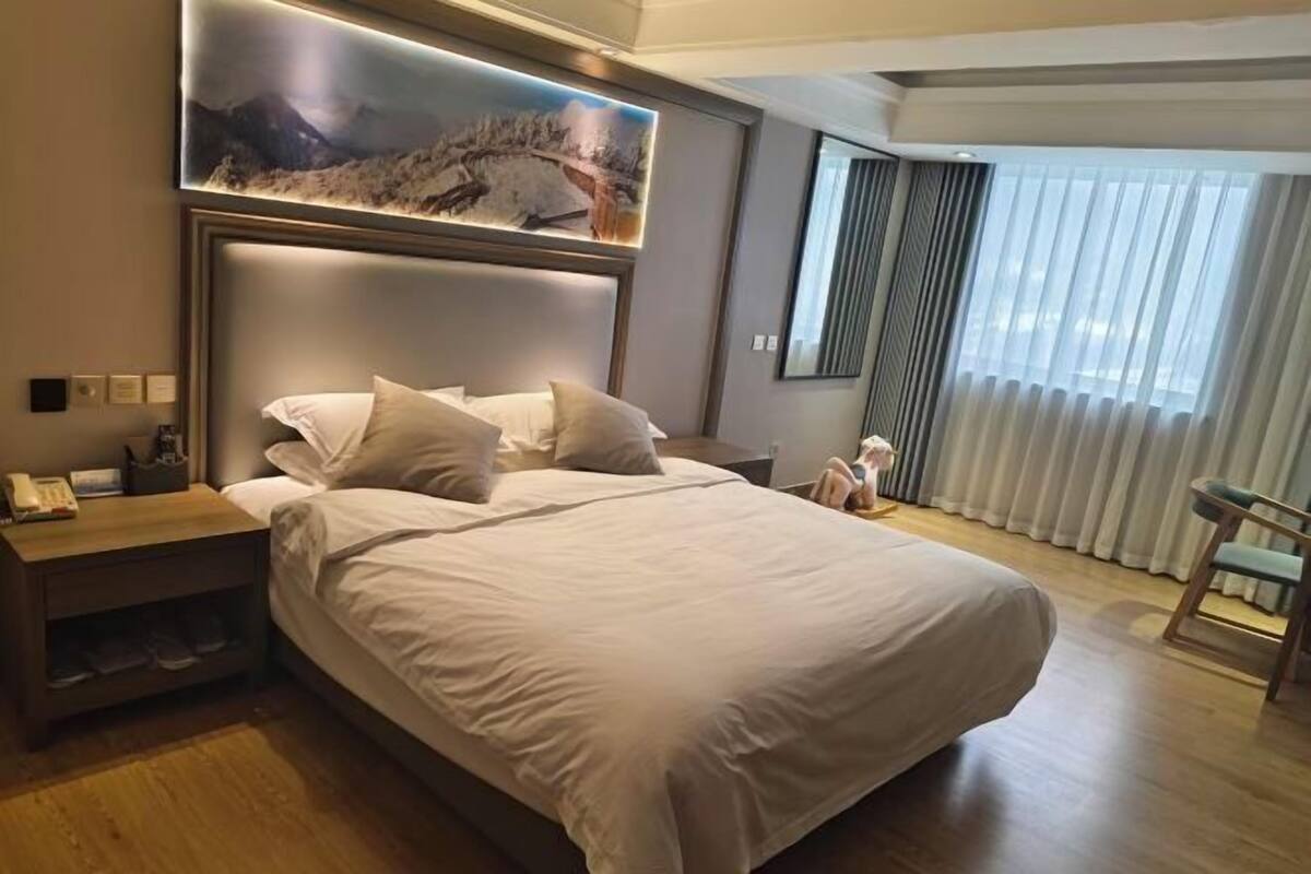 A spacious hotel room with a large bed, a bedside table with a lamp, a mounted artwork above the bed, and a view of the outdoors through sheer curtains.