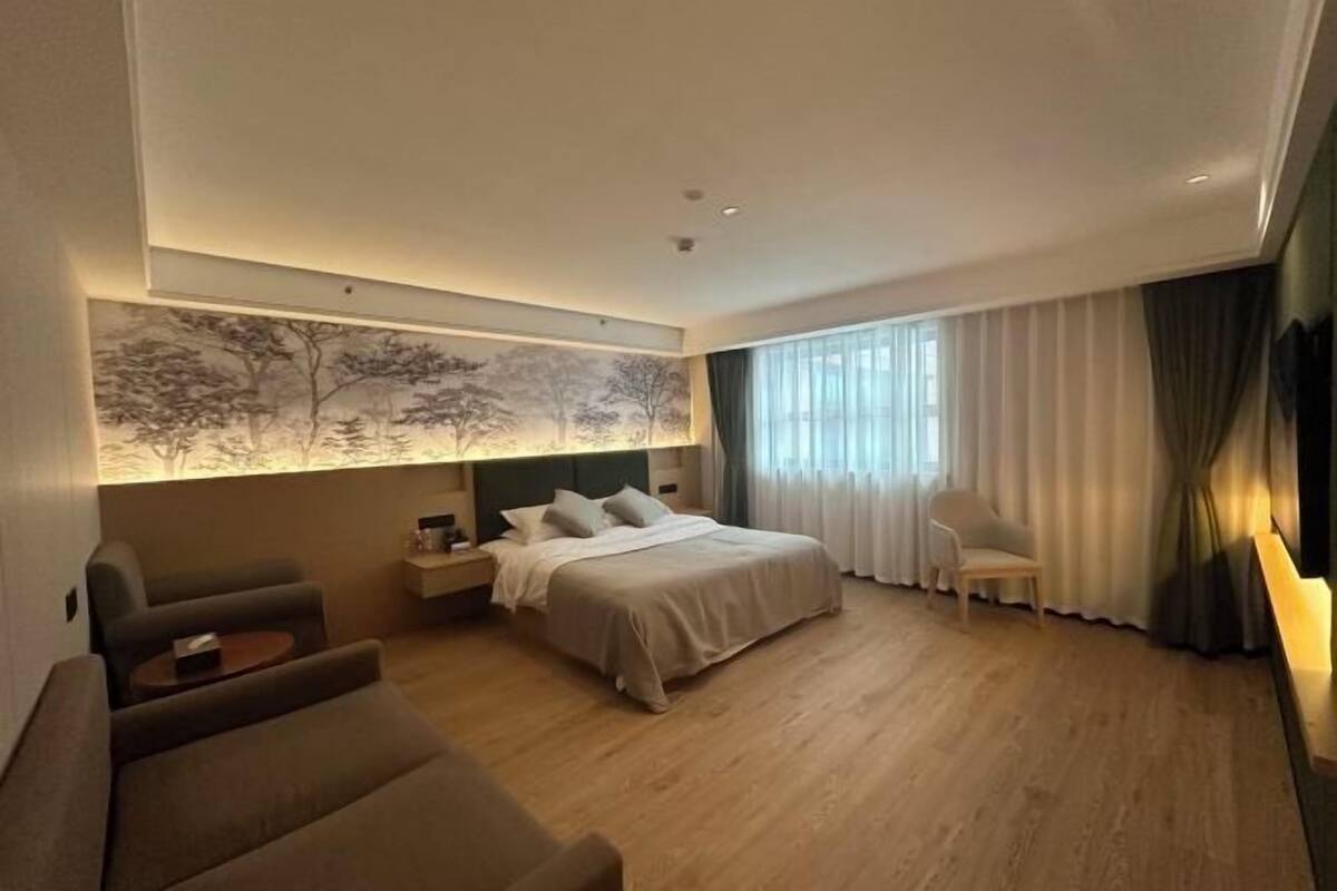 A modern hotel room with a large bed, a sofa, a desk, and a wall-mounted artwork.
