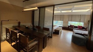 Un espace repas moderne avec une table et des chaises en bois, une grande fenêtre donnant sur un paysage verdoyant et une porte coulissante menant à un coin salon agrémenté de coussins.