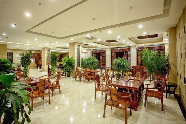 Un amplio hall de hotel con varias mesas y sillas de madera, rodeado de plantas en macetas.