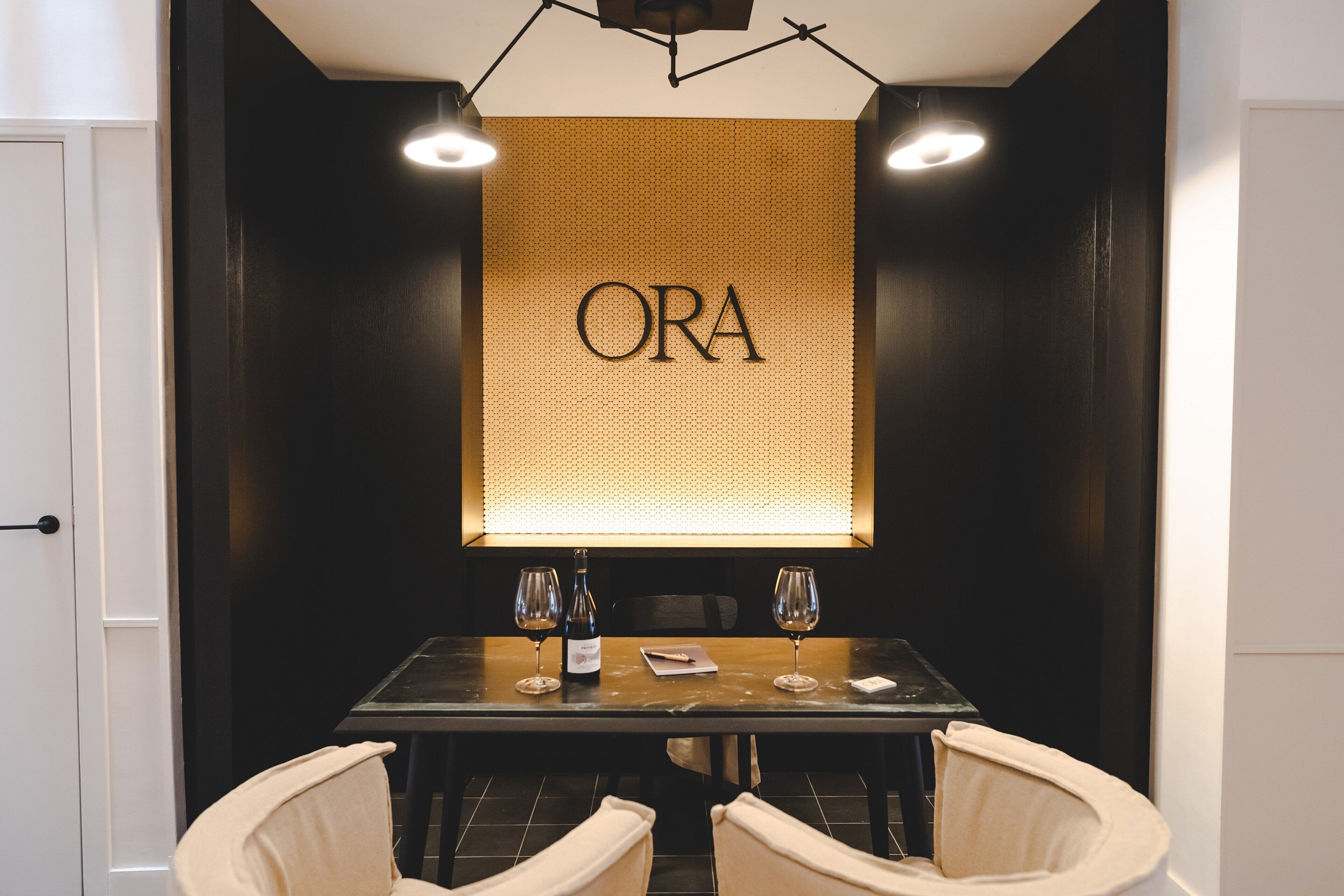 A dining area with a table set for two, two wine glasses, a bottle of wine, and a menu. The wall behind the table has the word "ORA" illuminated.