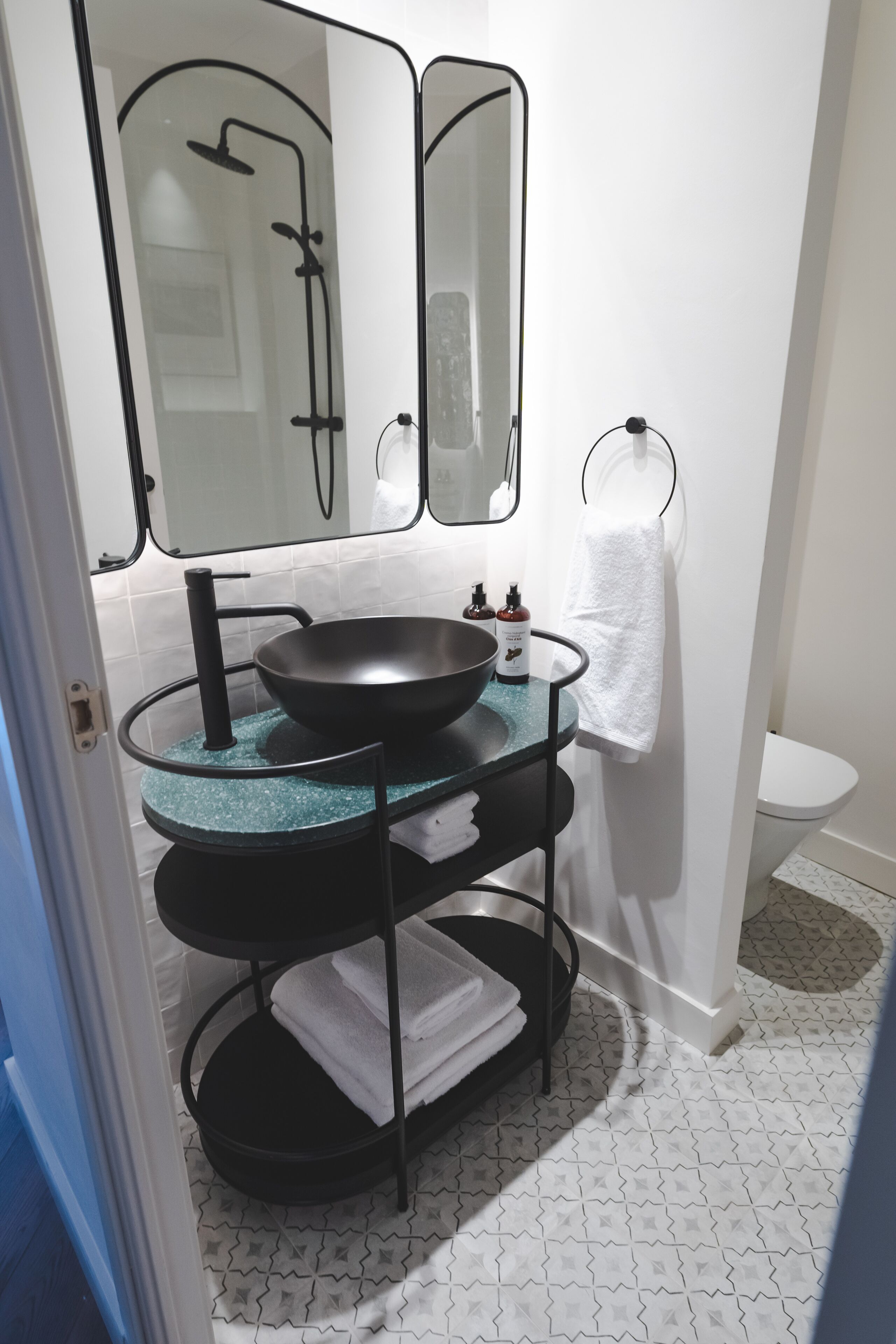 Un baño con un lavamanos negro, dos espejos, un perchero y un inodoro.