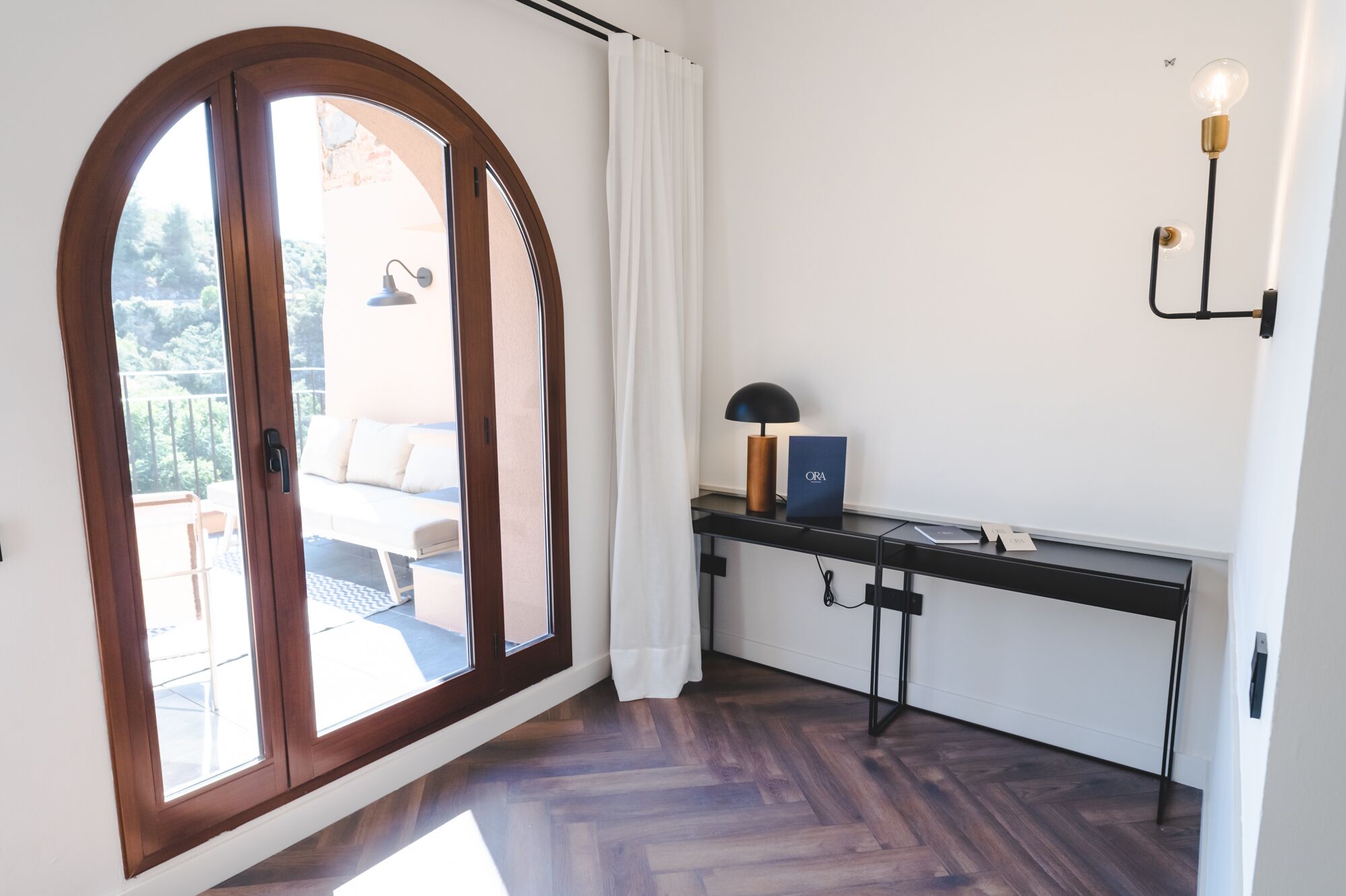 A room with a wooden floor, a desk with a lamp, and a view of an outdoor seating area through a large arched window.
