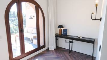 A room with a wooden floor, a desk with a lamp, and a view of an outdoor seating area through a large arched window.